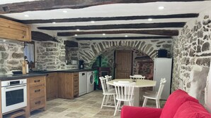 A rustic kitchen with stone walls, wooden cabinets, and a stone archway leading to a dining area with a red sofa and a round wooden table. - Entre Eaux et Roches - 5-person gite in character Ardèche house (Beaumont)