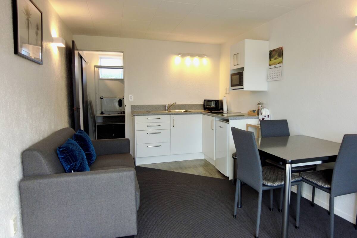 A compact kitchenette with a microwave, toaster, and sink. There is a dining table with four chairs and a sofa with a blue pillow.