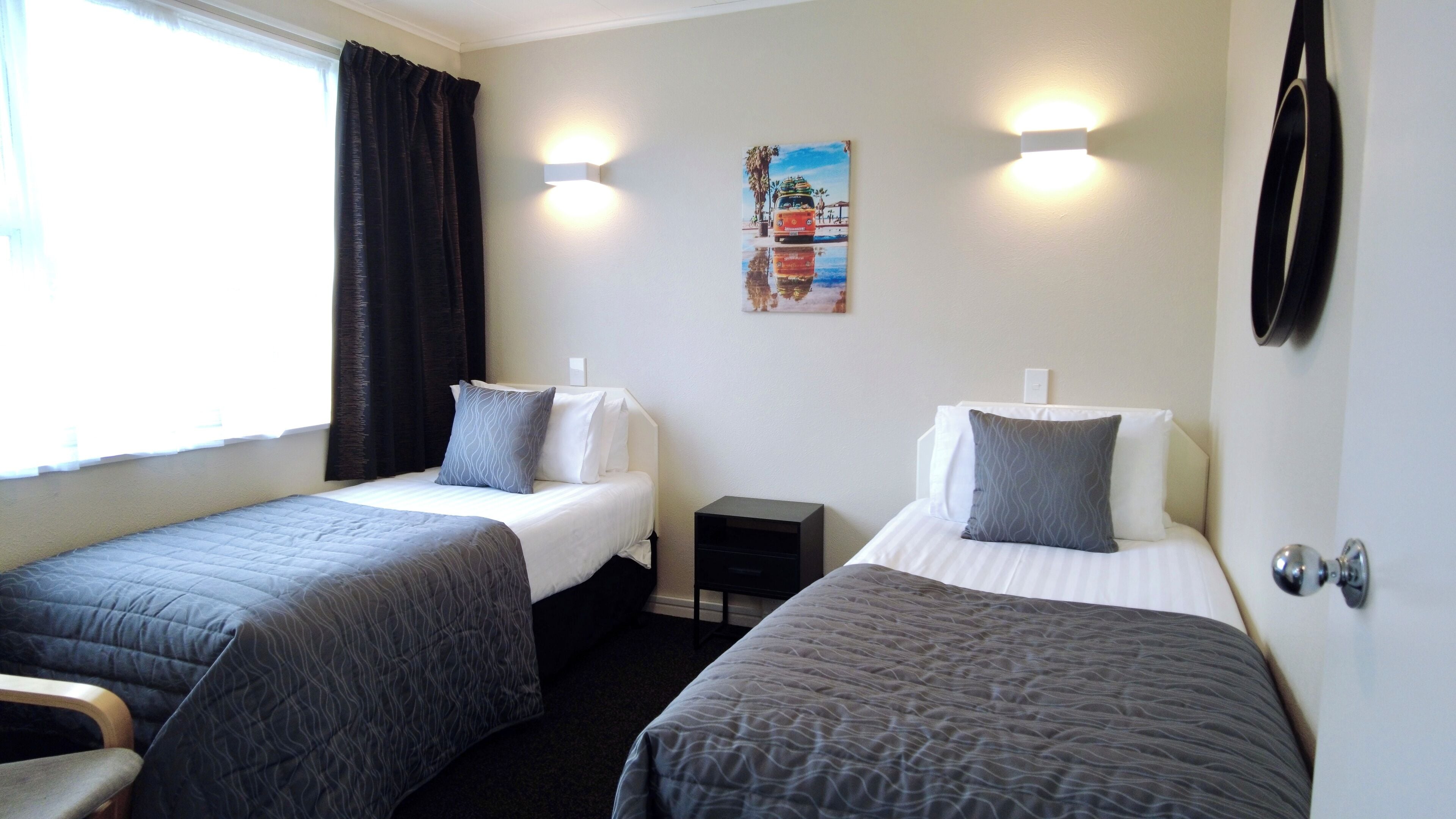 Habitación de hotel con dos camas individuales, una mesita de noche pequeña y un cuadro en la pared, además de un ventanal con cortinas.