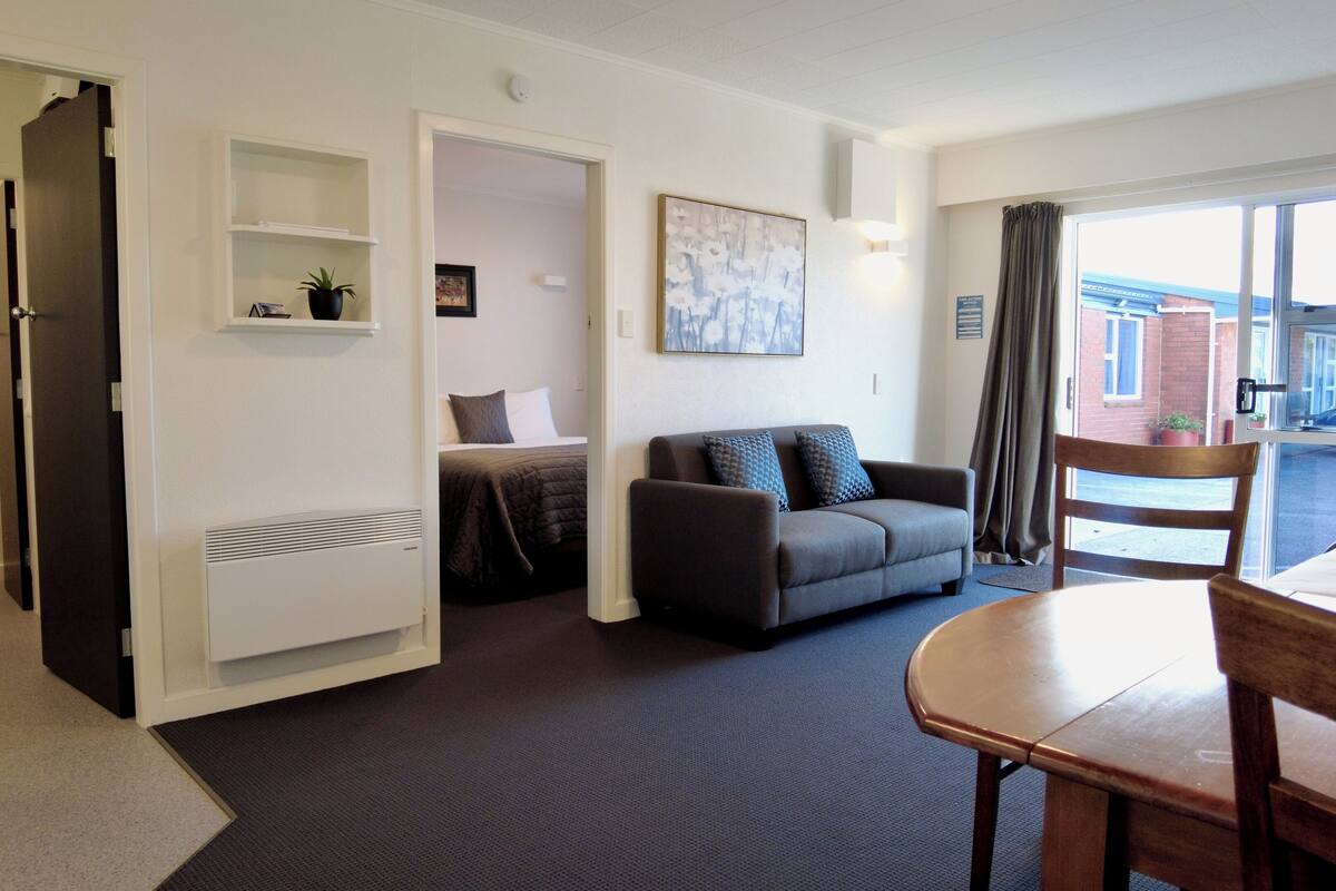 A modern hotel room with a sofa, a dining table, a small kitchen area, and a view of the outside.