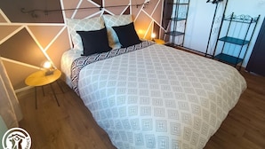 Ein Schlafzimmer mit einem bettenüberdeck in geometrischem Muster, zwei schwarzen Kissen, einem kleinen runden Tisch mit einer Lampe und einem Metallgestell mit darin hängenden Gegenständen.
