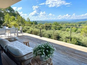 Una terraza de madera con un sofá y sillones de mimbre, una mesita con una planta en maceta y vistas a un paisaje boscoso.