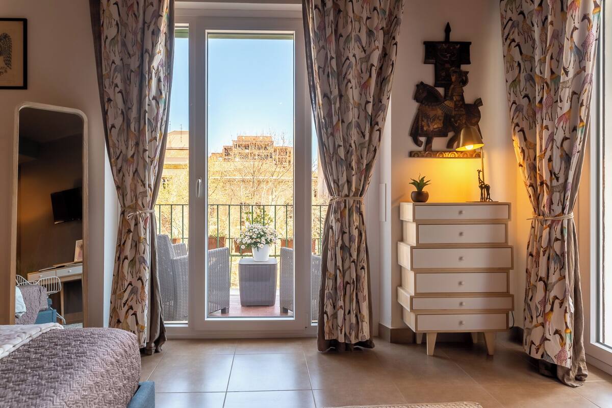 A bedroom with a bed, a dresser, a TV, and a balcony with wicker furniture and a potted plant.