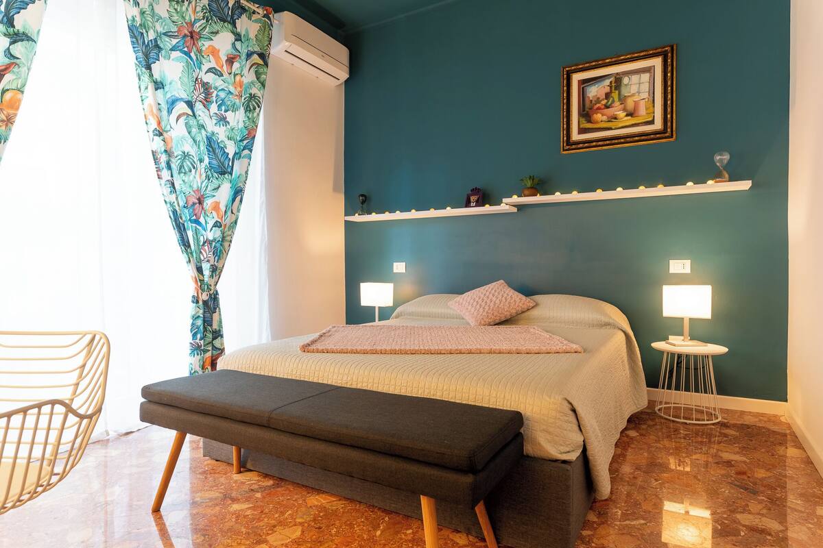 A bedroom with a bed, bedside lamps, a bench, a wall-mounted shelf, and a painting.