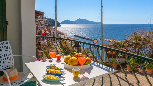 A balcony with a table set for a meal, overlooking a coastal view with mountains in the distance. - Estate4home - Bonview suite (Massa Lubrense)
