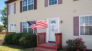 A two-story house with a front porch, a mailbox, and a small garden with a bush and flowers.