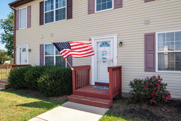 A two-story house with a front porch, a mailbox, and a small garden with a bush and flowers.