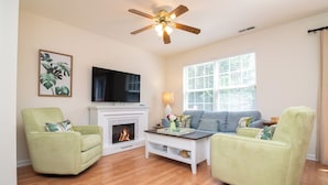 A living room with a fireplace, a sofa, two armchairs, a ceiling fan, and a television on the wall.
