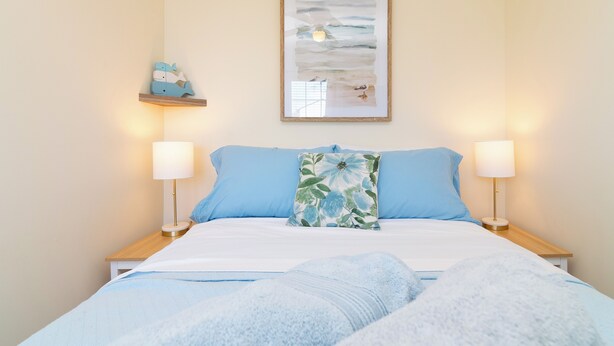 A neatly made bed with blue bedding, two bedside lamps, a framed artwork above the bed, and a small shelf with decorative items.