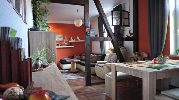 A cozy living room with a wooden floor, a sofa, a coffee table, and a dining table with a placemat. There is a bookshelf with books and decorative items, and a plant on the left side. - Midgard vacation apartment in Sagenharz - Family apartment, 2 bedrooms & WLAN (Thale)