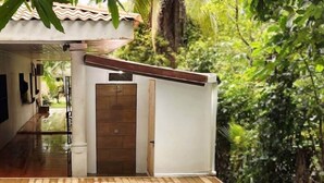 Un espace aménagé au bord de la piscine, avec une terrasse en bois, un mur blanc percé d’une porte et une vue sur une végétation luxuriante.