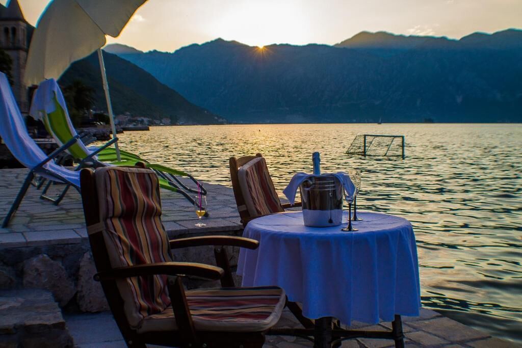 Ein Tisch mit Stühlen und einer Tischdecke am Seeufer, mit Blick auf einen stillen See und Berge im Hintergrund.
