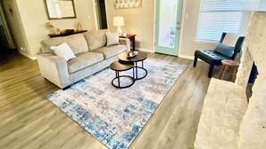 A living room with a beige sofa, a coffee table, and a patterned rug. There is a door with a light green frame and a window with blinds. - Round Rock - Austin Hideaway (Round Rock)