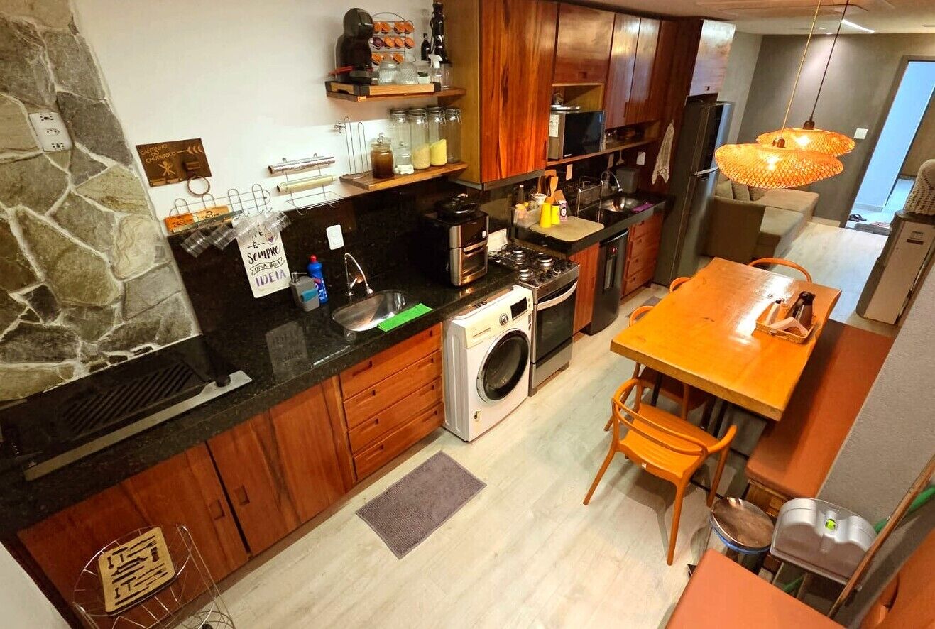 A modern kitchen with a stone accent wall, wooden cabinets, a built-in washing machine, and a dining area with orange chairs.