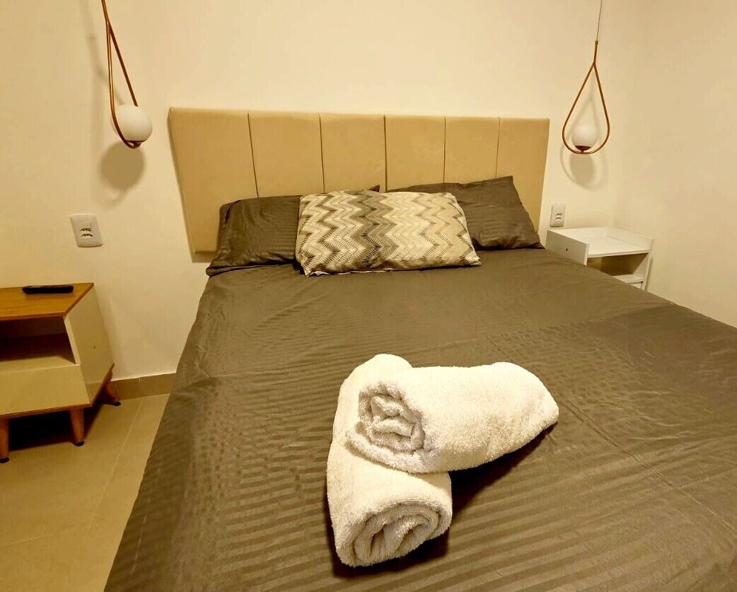 Una cama bien hecha con una toalla doblada, una mesita de noche y una lámpara de pared.