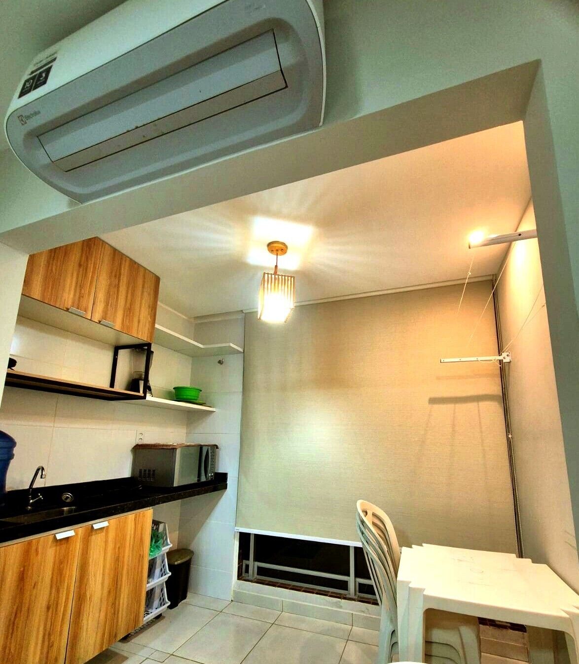 A compact kitchen with a built-in air conditioner, wooden cabinets, a black countertop, a white chair, and a hanging light fixture.