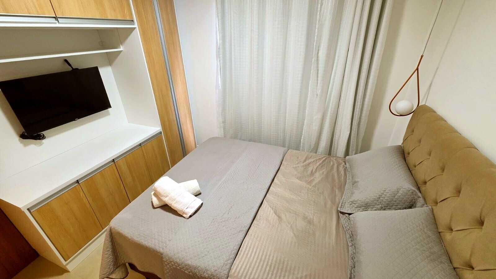 Een kleine, verzorgde kamer met een bed, een aan de muur bevestigde televisie en een gordijn.