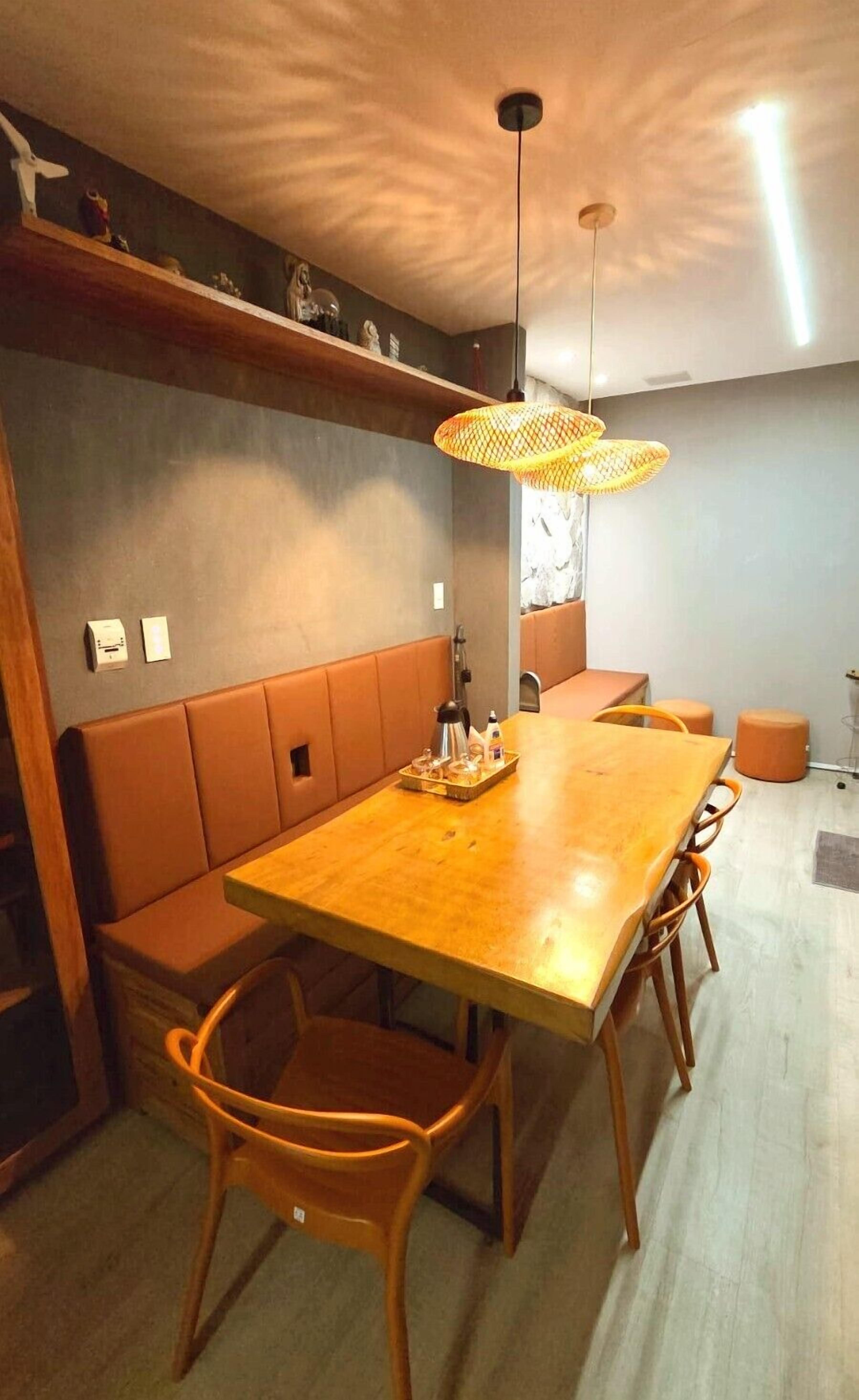 A dining area with a wooden table, cushioned bench seating, wicker pendant lights, and a wall-mounted shelf with decorative items.