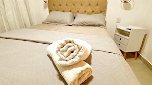 Een opgemaakt bed met een opgerolde handdoek, een nachtkastje met een lamp en een klein dressoir.