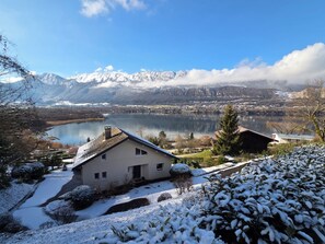 Une maison avec un toit en pente, un petit jardin et une vue sur un lac et des montagnes en arrière-plan.