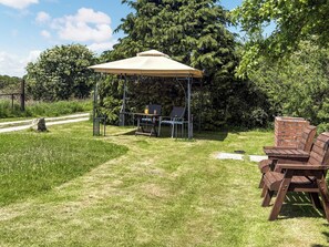 A covered outdoor seating area with a table and chairs, surrounded by greenery and trees.