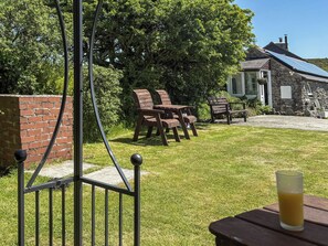 A grassy outdoor area with wooden chairs and a table, a stone building with a solar panel, and a metal fence.