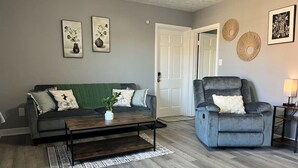 A living room with a grey sofa, a coffee table, and a grey armchair. There are two framed pictures on the wall and a door in the background. - Amazing Downtown Location! Quiet & Spacious! #C (Mount Vernon)