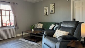 A living room with a grey sofa, a coffee table, and a radiator. There are two framed pictures on the wall and a window with curtains. - Amazing Downtown Location! Quiet & Spacious! #C (Mount Vernon)