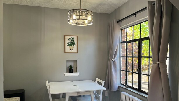 A dining area with a white table and chairs, a pendant light, a framed plant, a small shelf with a plant, a window with curtains, and a radiator. - Amazing Downtown Location! Quiet & Spacious! #C (Mount Vernon)