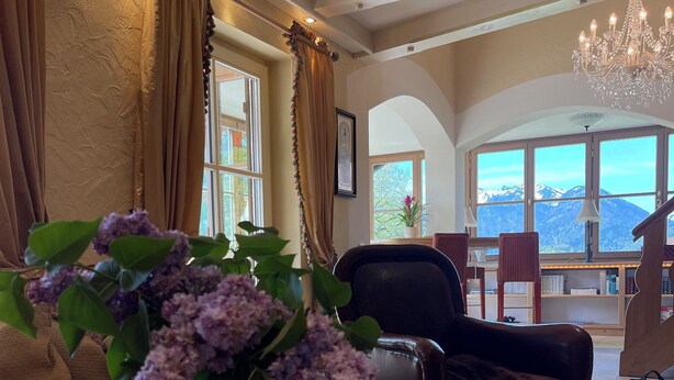 A cozy living room with a wooden coffee table, a vase with flowers, a bookshelf, and a view of mountains through large windows.