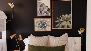 A modern bedroom with a large bed, striped pillows, and a green throw. There are framed botanical prints on the wall and a bedside lamp. - Lucas Oil Stadium Sanctuary! Hot Tubs! Games! (Indianapolis)