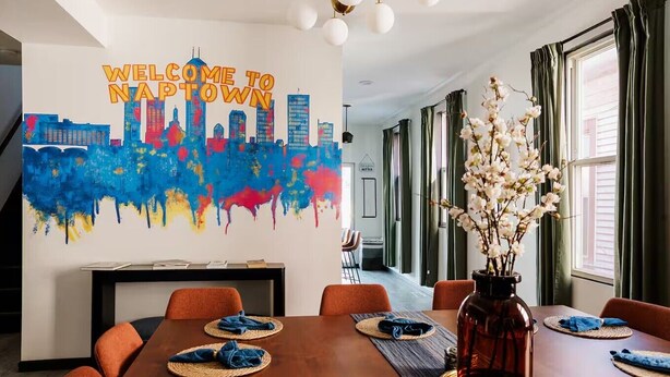 A modern hotel lobby with a "Welcome to Nap Town" mural, a wooden reception desk, and a dining area with orange chairs and blue napkins. - Lucas Oil Stadium Sanctuary! Hot Tubs! Games! (Indianapolis)