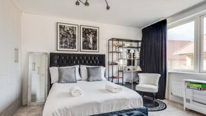 Un dormitorio con una cama grande, una mesita de noche, una silla, una estantería y una ventana con cortinas.