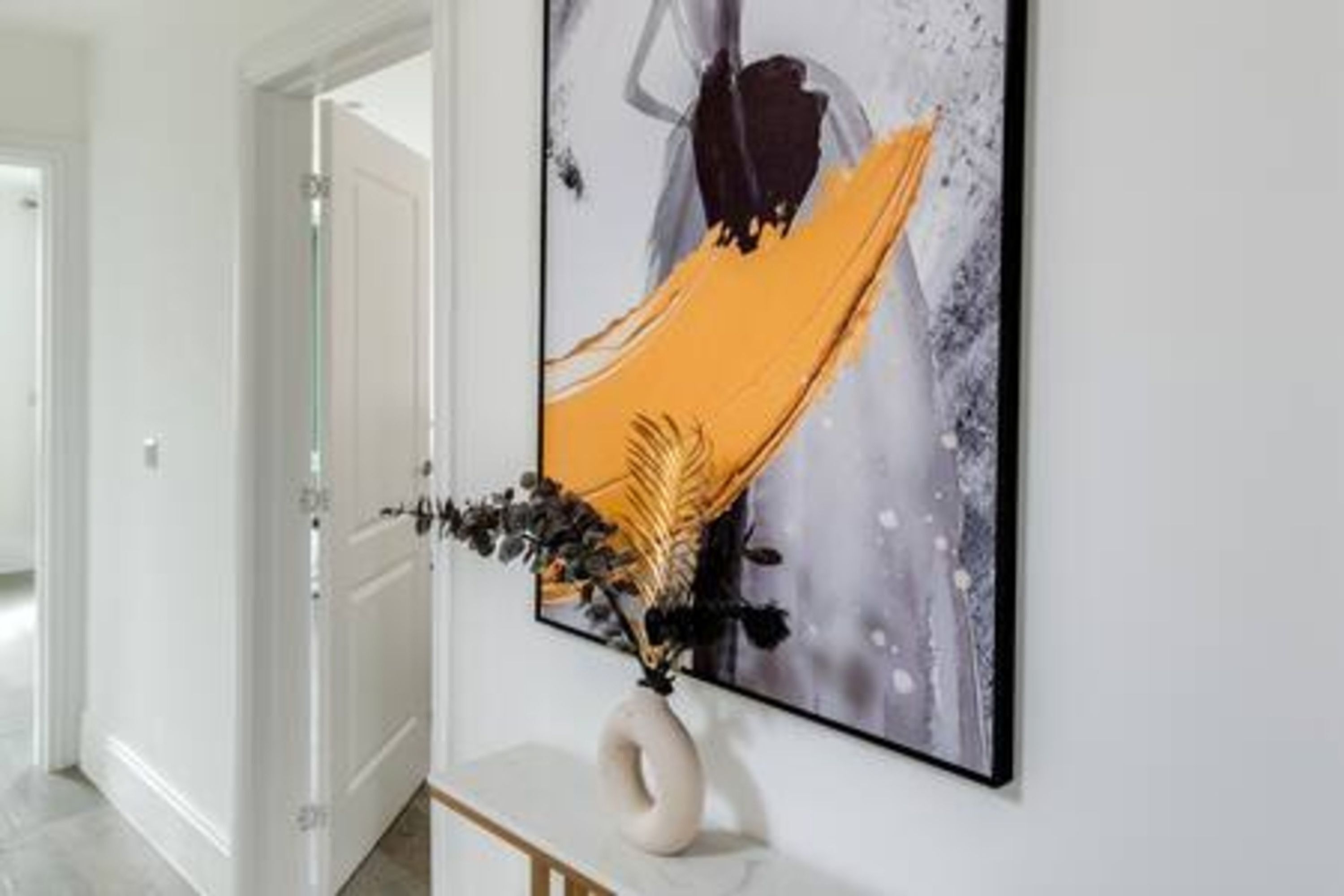 A hallway with a framed abstract painting on the wall, a white vase with a plant, and a wooden console table.