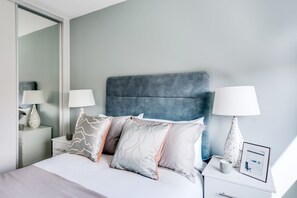 A modern bedroom with a large bed, grey and white bedding, two bedside tables with lamps, and a large mirror.