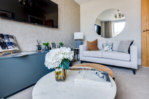 A modern living room with a white sofa, a round coffee table, a vase with white flowers, a flat-screen TV mounted on the wall, and a round mirror above the sofa.