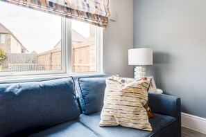 A room with a blue sofa, a patterned pillow, a white lamp, and a window with a view of a wooden fence and houses. - Stylish 4-br House - Free Parking Nice Garden (Oxford)