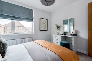 Ein Schlafzimmer mit Bett, Schminktisch, Spiegel, Fenster mit Blick auf Häuser und Heizkörper.