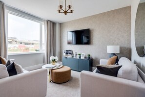 Ein modernes Hotelzimmer mit großem Fenster, einem flachbildfernseher an der Wand, einem runden Hocker und einem grauen Sofa.