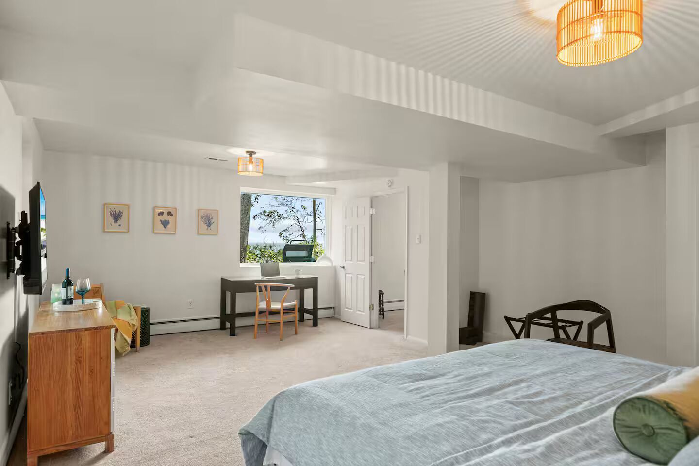 Une chambre à coucher avec un lit, un bureau et une chaise. Il y a une fenêtre donnant sur des arbres et une porte menant à une autre pièce.