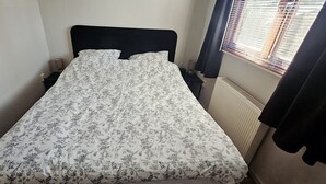 Un lit simple avec une couvre-lit à motifs floraux, une tête de lit noire, une table de chevet, une lampe et une fenêtre avec des stores.