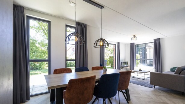 Un comedor moderno con una mesa y sillas de madera, ventanas amplias con cortinas y un sofá al fondo.