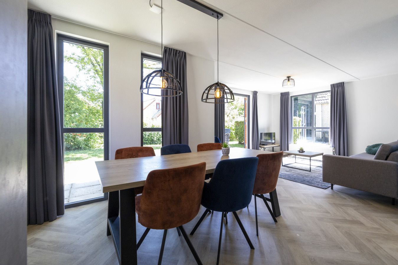 A modern dining area with a wooden table and chairs, large windows with curtains, and a sofa in the background.