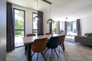 A modern dining area with a wooden table and chairs, large windows with curtains, and a sofa in the background.