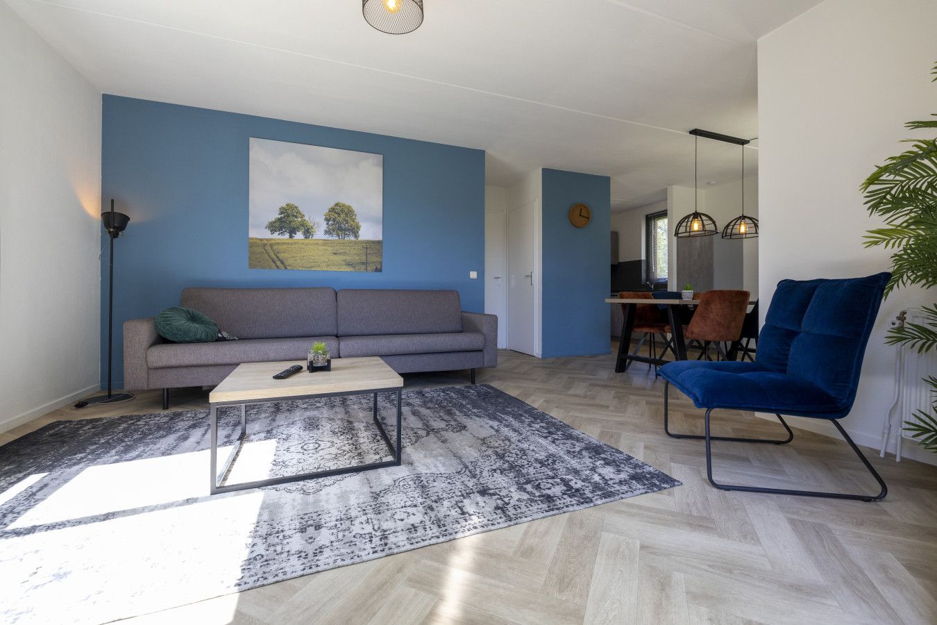 Uma sala de estar moderna com um sofá cinza, uma mesa de centro e uma poltrona azul. Há um quadro na parede e uma planta no canto.