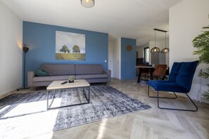 En moderne stue med en grå sofa, et sofabord og en blå lænestol. Et maleri hænger på væggen, og der står en plante i hjørnet.