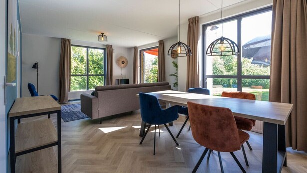 A modern living room with a sofa, dining table, and chairs. There are large windows with curtains, a hanging light fixture, and a view of greenery outside.