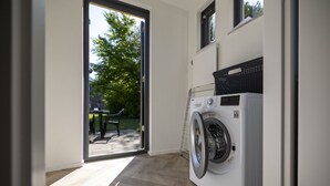A laundry room with a front-loading washing machine, a drying rack, and a view of the outdoors through a glass door.