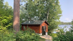 A small cabin with a chimney, surrounded by dense greenery and a gravel path leading to it. - Lake Vermilion Cabin on a quiet bay with sauna & fire pit for grilling. (Cook)