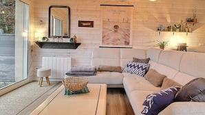 Una sala de estar moderna con un sofá modular, una mesa de centro y un ventanal grande. Hay un cartel en la pared y una pequeña estantería con plantas.